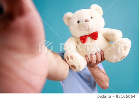 Portrait of unknown bearded man making selfie or broadcasting livestream, point of view photo, hiding his face behind soft teddy bear. Indoor studio shot isolated on blue background. 95166064