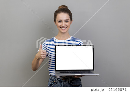 Happy smiling satisfied woman wearing striped T-shirt holding laptop with empty blank display and showing thumbs up, pleased with online application. Indoor studio shot isolated on gray background. 95166071