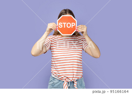 Portrait of unknown woman wearing T-shirt holding and hiding her face with red stop sign, holding red traffic sign, attention to safe road crossing. Indoor studio shot isolated on purple background. Portrait of unknown woman wearing T-shirt holding and hiding her face with red stop sign, holding red traffic sign, attention to safe road crossing. Indoor studio shot isolated on purple background. 95166164