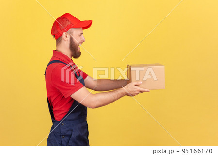 Profile portrait of positive deliveryman holding cardboard parcel, delivering order door-to-door, shipment and cargo transportation service. Indoor studio shot isolated on yellow background. Profile portrait of positive deliveryman holding cardboard parcel, delivering order door-to-door, shipment and cargo transportation service. Indoor studio shot isolated on yellow background. 95166170