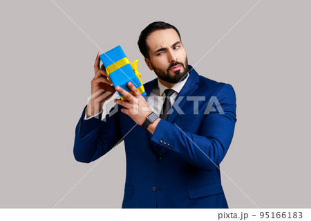 Portrait of curious bearded businessman holding and shaking wrapped present box, being interested what inside, wearing official style suit. Indoor studio shot isolated on gray background. 95166183