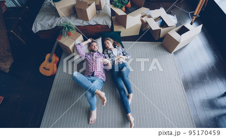Top view of loving young couple lying on bedroom floor in new apartment and talking. Guitar, packed things in carton boxes, double bed and modern carpet are visible. Top view of loving young couple lying on bedroom floor in new apartment and talking. Guitar, packed things in carton boxes, double bed and modern carpet are visible. 95170459