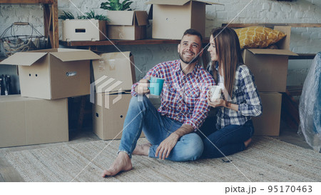 Girlfriend and boyfriend are talking making plans sitting on floor of new purchased apartment, looking around and holding mugs. Relationship, people and relocation concept. 95170463