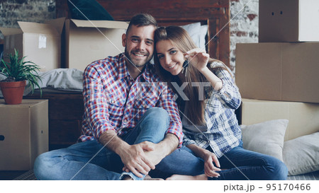Portrait of happy young couple sitting together in new flat, holding keys and looking at camera. Nice loft style interior, carton boxes and double bed are visible. Portrait of happy young couple sitting together in new flat, holding keys and looking at camera. Nice loft style interior, carton boxes and double bed are visible. 95170466