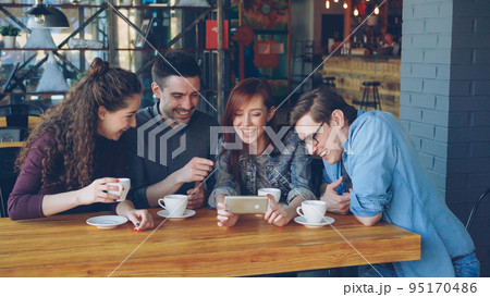 Happy good-looking students are watching funny video, discussing it and laughing during meeting in cafe. Cheerful people, modern technology and eating out concept. Happy good-looking students are watching funny video, discussing it and laughing during meeting in cafe. Cheerful people, modern technology and eating out concept. 95170486