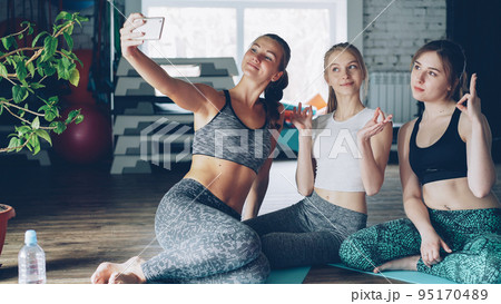 Attractive young women are taking selfie with smartphone after yoga class in light modern gym in wellness center. Women are wearing trendy sports clothes. Sports equipment in background. 95170489
