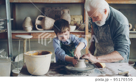 Concentrated little boy is learning to work with clay on professional throwing-wheel in pottery class in traditional workshop. His teacher senior experienced man is helping him. 95170550