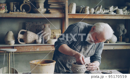 Experienced craftsman bearded man is making ear for ceramic bowl and fixing it on clayware while working in cozy workplace. Pottery, profession and hobby concept. Experienced craftsman bearded man is making ear for ceramic bowl and fixing it on clayware while working in cozy workplace. Pottery, profession and hobby concept. 95170559