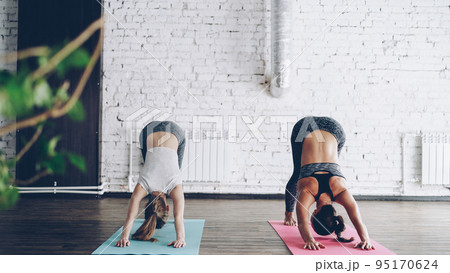 Young blond girl is enjoying individual yoga practice with friendly female instructor in light studio. Women are doing sequence of asanas on bright mats. Friendly relaxing atmosphere. 95170624