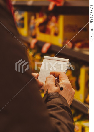 Female hands holding paper notebook of shopping list, blurred shelves on background. Partial view of woman doing shopping in supermarket. Concept of consumerism Close up of woman making notes in 95172373