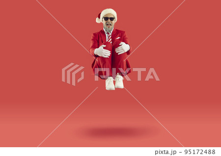 Old cheerful grey-haired man wearing Santa style is sitting levitating in air on red background. Old cheerful grey-haired man wearing Santa style is sitting levitating in air on red background. 95172488