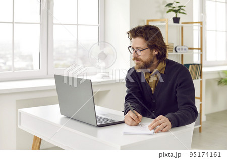 Redhead Caucasian man entrepreneur looks at laptop and making notes in notebook sits at office desk 95174161
