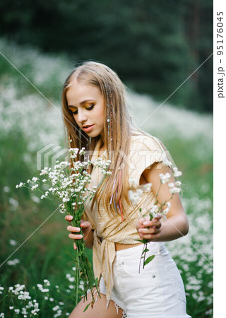 Beautiful young woman picking white wildflowers on the background of a forest landscape in summer. Portrait of a gentle happy woman in a wild field, enjoying nature. Natural Beauty Model 95176045