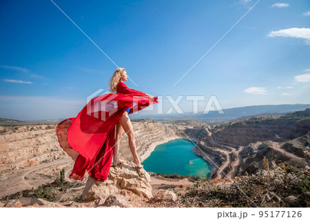 Side view of a beautiful sensual woman in a red long dress posing on a rock high above the lake in the afternoon. Against the background of the blue sky and the lake in the form of a heart 95177126