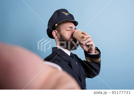 Pilot drinking coffee to go first person view, taking selfie on front camera, fpv. Middle aged airlane captain in uniform holding take away tea paper cup pox, making photo on mobile phone 95177731
