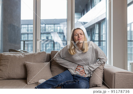 A young woman is sitting on the couch at home, holding on to a stomach that hurts, screams in pain, feels pain, is sick, feels bad A young woman is sitting on the couch at home, holding on to a stomach that hurts, screams in pain, feels pain, is sick, feels bad 95178736