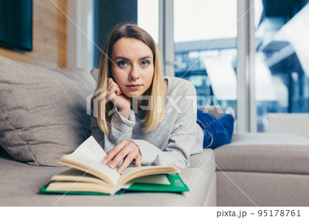 Portrait of a young beautiful woman reading a book, lying on the couch, resting, looking at the camera, smiling Portrait of a young beautiful woman reading a book, lying on the couch, resting, looking at the camera, smiling 95178761