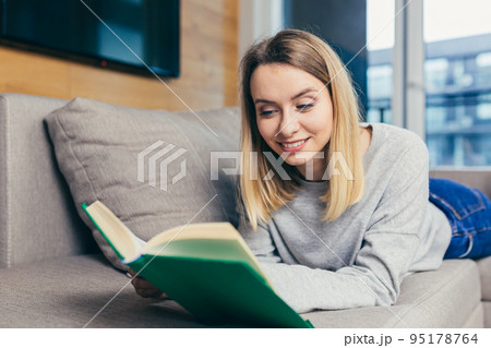 Portrait of a young beautiful woman reading a book, lying on the couch, resting, looking at the camera, smiling Portrait of a young beautiful woman reading a book, lying on the couch, resting, looking at the camera, smiling 95178764