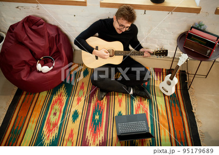 Aerial view of young man siiting on floor at home and leaning to play guitar. Hobbies and leisure activities. Remote training, online classes Aerial view of young man siiting on floor at home and leaning to play guitar. Hobbies and leisure activities. Remote training, online classes 95179689