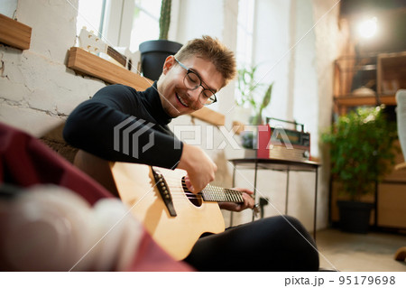 Young man siiting on floor at home and leaning to play guitar. Hobbies and leisure activities. Remote training, online classes Young man siiting on floor at home and leaning to play guitar. Hobbies and leisure activities. Remote training, online classes 95179698