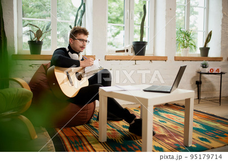 Young man siiting on floor at home and leaning to play guitar. Hobbies and leisure activities. Remote training, online classes Young man siiting on floor at home and leaning to play guitar. Hobbies and leisure activities. Remote training, online classes 95179714