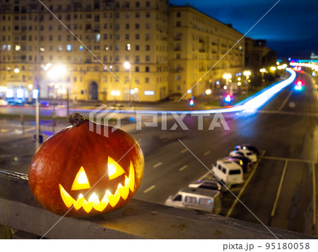 Halloween pumpkin on the background of the night city. Buildings and streaks of light from cars passing along the road. Halloween pumpkin on the background of the night city. Buildings and streaks of light from cars passing along the road. 95180058