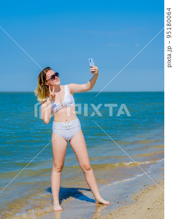 Beautiful young girl on the sea beach near the water posing and taking a selfie on a smartphone. Girl in a white swimsuit and sunglasses. Summer sunny day. Sea vacation concept. Copy space 95180064
