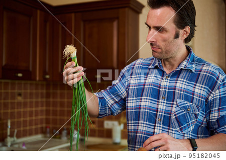 Handsome middle-aged Caucasian man in blue checkered casual shirt, standing in the kitchen at home, sorting fresh organic vegetables and groceries while unpacking packet after grocery shopping Handsome middle-aged Caucasian man in blue checkered casual shirt, standing in the kitchen at home, sorting fresh organic vegetables and groceries while unpacking packet after grocery shopping 95182045