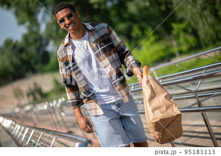 Young dark-skinned man in plaid shirt walking with a paper bag in hands Young dark-skinned man in plaid shirt walking with a paper bag in hands 95183113
