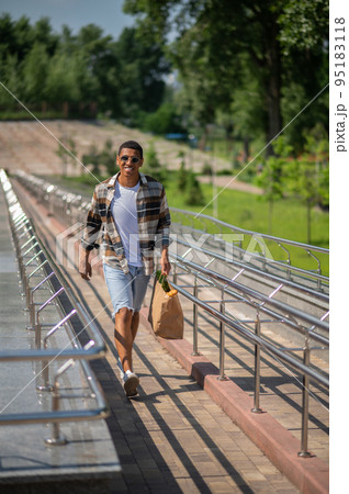Young dark-skinned man in plaid shirt walking with a paper bag in hands 95183118