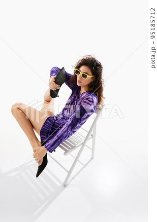 Portrait of young beautiful woman sitting on chair, posing in purple short dress over white background. Leg stretching 95185712