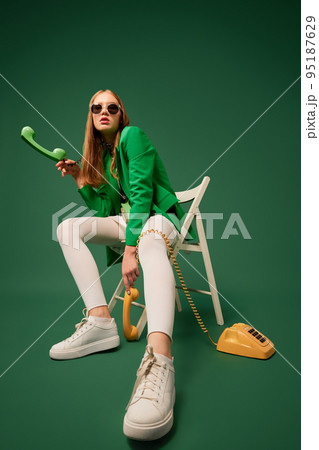 Call your parents. Creative portrait of young without emotions girl in green jacket sitting on chair with retro phone isolated over green background. Call your parents. Creative portrait of young without emotions girl in green jacket sitting on chair with retro phone isolated over green background. 95187629