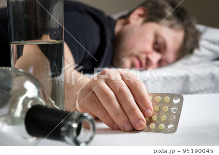 A man lies on a bed after an alcohol party, holding hangover pills in his hand A man lies on a bed after an alcohol party, holding hangover pills in his hand 95190045