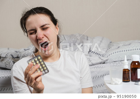 A woman with insomnia sits by the bed and holds pills for healthy sleep and yawns. 95190050