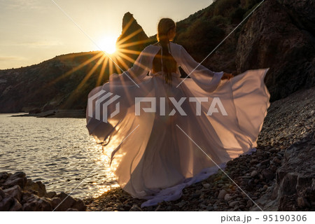 A mysterious female silhouette with long braids stands on the sea beach with mountain views, Sunset rays shine on a woman. Throws up a long white dress, a divine sunset. 95190306