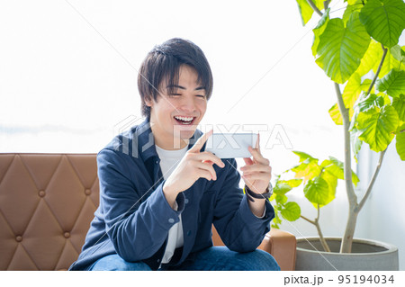 スマホで動画視聴する20代男性-リビングのソファーに座りリラックス スマホで動画視聴する20代男性-リビングのソファーに座りリラックス 95194034