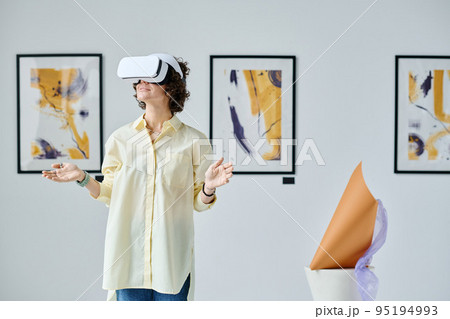 Young woman using virtual reality glasses standing at gallery of modern art during exhibition Young woman using virtual reality glasses standing at gallery of modern art during exhibition 95194993