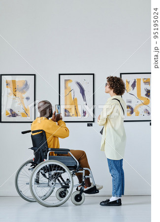 Rear view of African man in wheelchair making photo on mobile phone while visiting art gallery together with woman 95195024