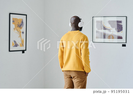 Rear view of African young man in wireless headphones looking at modern art and listening to guide 95195031