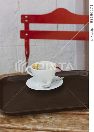 A white coffee cup on a saucer stands on a tray on a wooden table on the terrace of a cafe, a coffee shop in an urban urban environment. Conceptual vertical photo with a hot beverage drink. Red chair. A white coffee cup on a saucer stands on a tray on a wooden table on the terrace of a cafe, a coffee shop in an urban urban environment. Conceptual vertical photo with a hot beverage drink. Red chair. 95196271