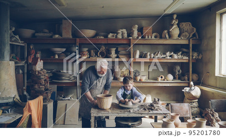 Skilled child is forming pot on potter's wheel while working with his retired grandfather in his figuline home studio. Professional equipment and beautiful ceramic things in background. 95201780