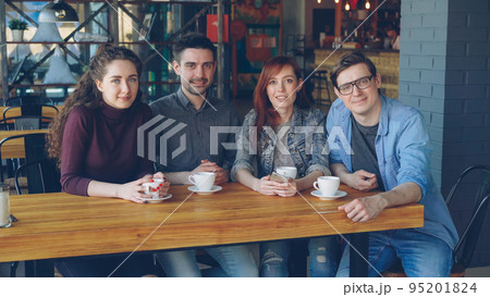 Portrait of four smiling young people friends in casual clothes sitting at table in spacious cafe with tea cups and looking at camera. Eating out and friendship concept. 95201824