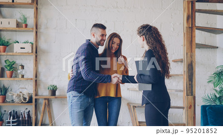 Friendly realtor is giving keys to young couple buyers of new house, happy spouses are hugging , smiling man is shaking hands with broker making deal. 95201852
