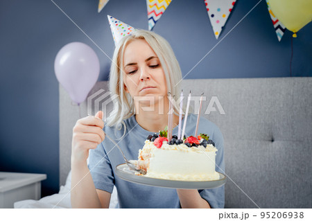 Sad, upset woman in pajama and party cap thinking eat or not to eat birthday cake sitting on the bed in decorated bedroom. Celebrates birthday alone concept. Selective focus Sad, upset woman in pajama and party cap thinking eat or not to eat birthday cake sitting on the bed in decorated bedroom. Celebrates birthday alone concept. Selective focus 95206938
