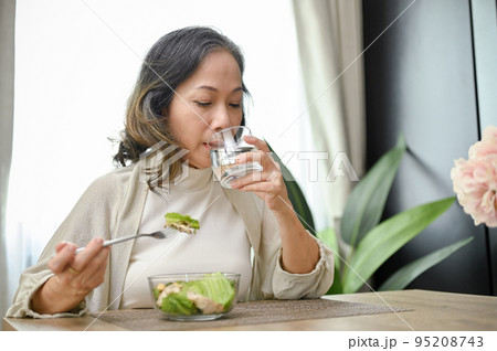 Healthy Asian-aged eating salad and drinking water in her dining room. 95208743