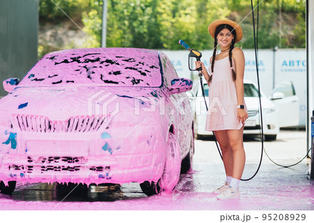 Woman with hose stands by car covered in pink foam Woman with hose stands by car covered in pink foam 95208929