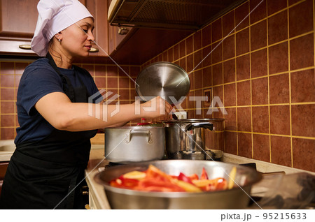 Charming housewife woman in a white chef's hat and black apron, stirs boiling tomato juice in a saucepan with a kitchen spoon while making homemade passata. Step-by-step recipe canning vegetables Charming housewife woman in a white chef's hat and black apron, stirs boiling tomato juice in a saucepan with a kitchen spoon while making homemade passata. Step-by-step recipe canning vegetables 95215633