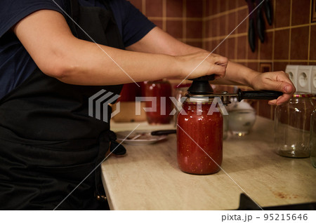 Close-up. Selective focus on a chef's hands using seamer or special seaming key, close lids of jars with freshly canned tomato sauce, juice or passata. Canning homegrown organic tomatoes for winter Close-up. Selective focus on a chef's hands using seamer or special seaming key, close lids of jars with freshly canned tomato sauce, juice or passata. Canning homegrown organic tomatoes for winter 95215646