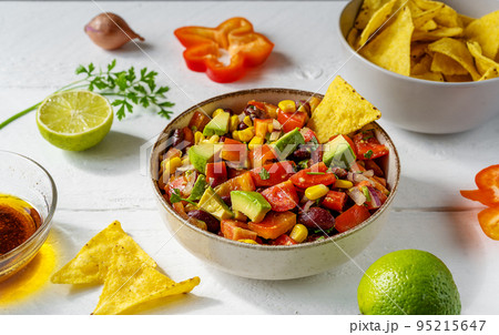 Cowboy caviar is traditional Mexican vegetable salad in bowl with ingredients and nachos on white wooden table with ingredients Cowboy caviar is traditional Mexican vegetable salad in bowl with ingredients and nachos on white wooden table with ingredients 95215647