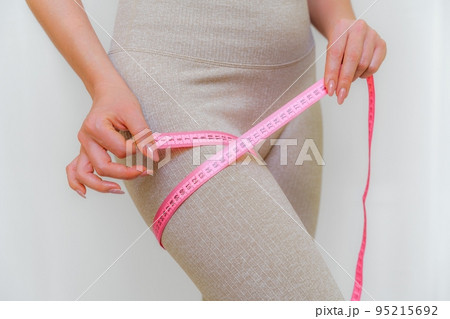 Cropped view of slim woman measuring her leg with tape measure at home, close-up. An unrecognizable European woman checks the result of a weight loss diet or liposuction indoors. Healthy lifestyle. Cropped view of slim woman measuring her leg with tape measure at home, close-up. An unrecognizable European woman checks the result of a weight loss diet or liposuction indoors. Healthy lifestyle. 95215692